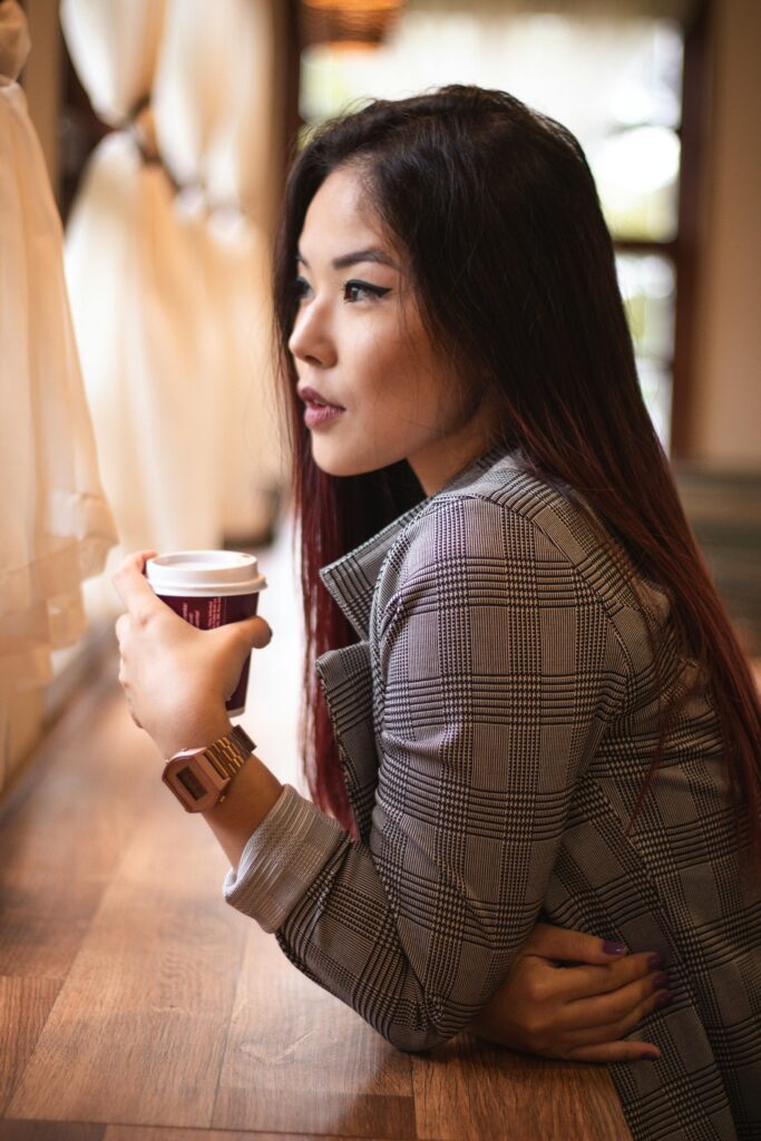 Stylish Asian woman in plaid coat sipping coffee by a sunlit window indoors.